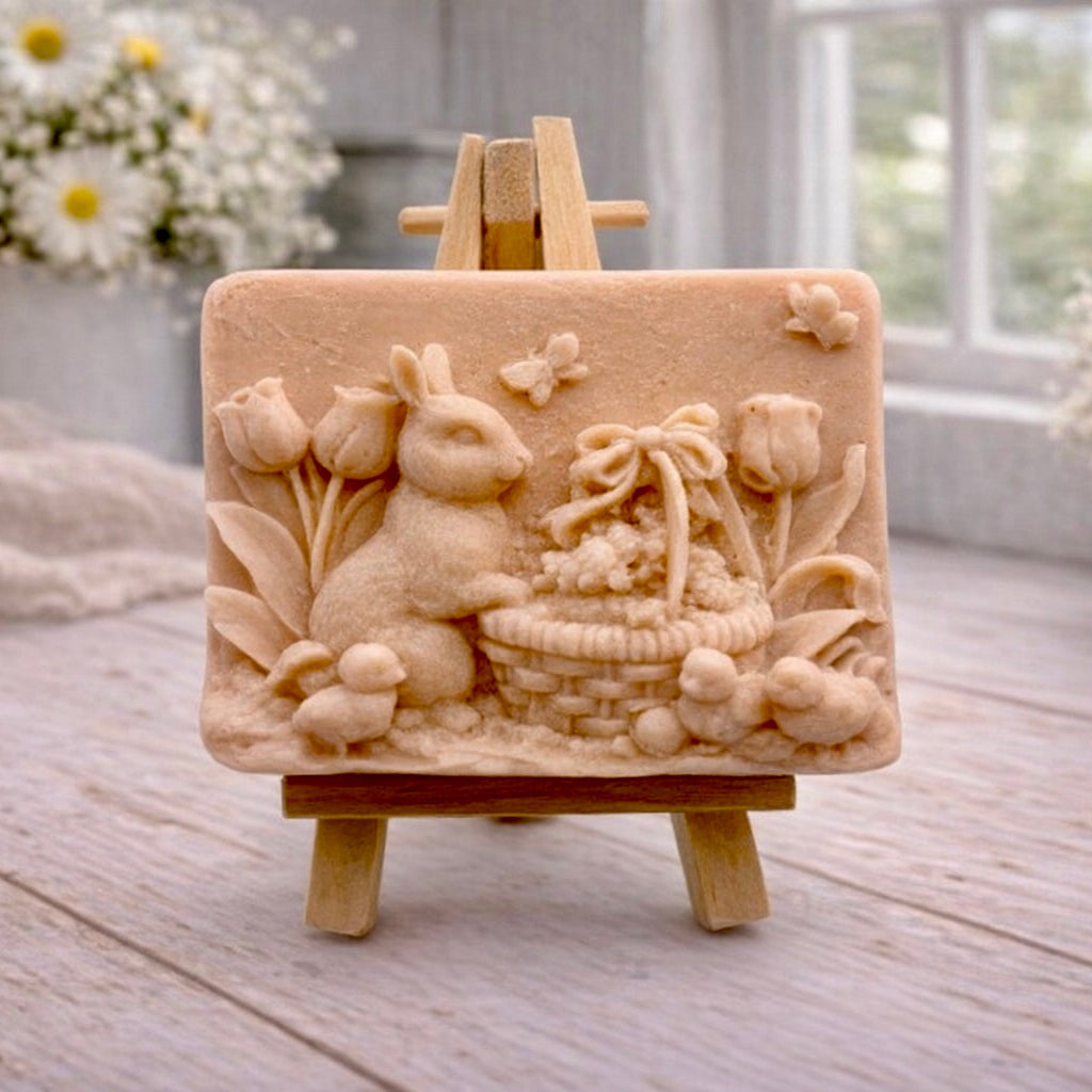 A rectangular handmade vegan soap in a sophisticated tone, featuring a highly detailed relief scene of an Easter bunny standing next to a basket of eggs, surrounded by blooming tulips and small birds, displayed on a wooden easel.