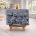 A rectangular handmade vegan soap in a sophisticated slate-grey tone, featuring a highly detailed relief scene of an Easter bunny standing next to a basket of eggs, surrounded by blooming tulips and small birds, displayed on a wooden easel.