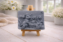 Load image into Gallery viewer, A rectangular handmade vegan soap in a sophisticated slate-grey tone, featuring a highly detailed relief scene of an Easter bunny standing next to a basket of eggs, surrounded by blooming tulips and small birds, displayed on a wooden easel.

