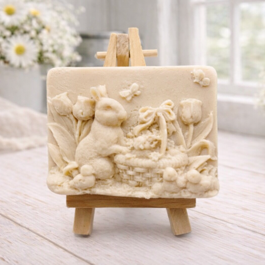 A rectangular handmade vegan soap in a sophisticated slate-grey tone, featuring a highly detailed relief scene of an Easter bunny standing next to a basket of eggs, surrounded by blooming tulips and small birds, displayed on a wooden easel.