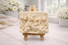 Load image into Gallery viewer, A rectangular handmade vegan soap in a sophisticated slate-grey tone, featuring a highly detailed relief scene of an Easter bunny standing next to a basket of eggs, surrounded by blooming tulips and small birds, displayed on a wooden easel.
