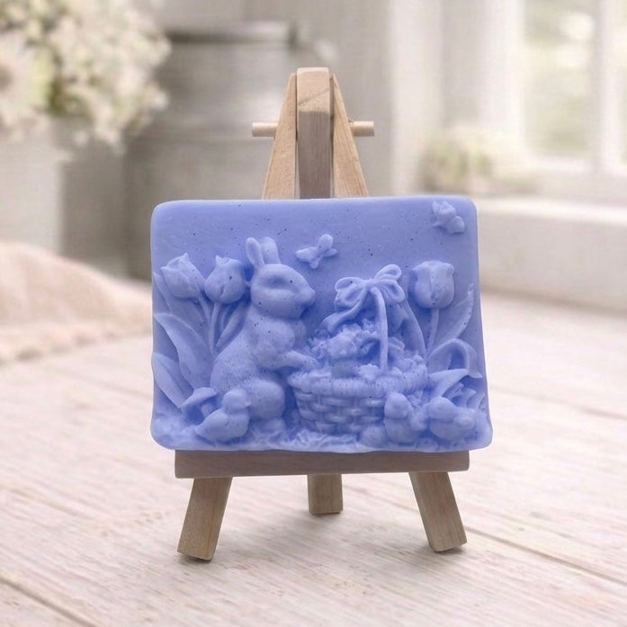 A rectangular handmade vegan soap in a sophisticated tone, featuring a highly detailed relief scene of an Easter bunny standing next to a basket of eggs, surrounded by blooming tulips and small birds, displayed on a wooden easel.
