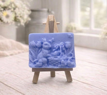 Load image into Gallery viewer, A rectangular handmade vegan soap in a sophisticated tone, featuring a highly detailed relief scene of an Easter bunny standing next to a basket of eggs, surrounded by blooming tulips and small birds, displayed on a wooden easel.
