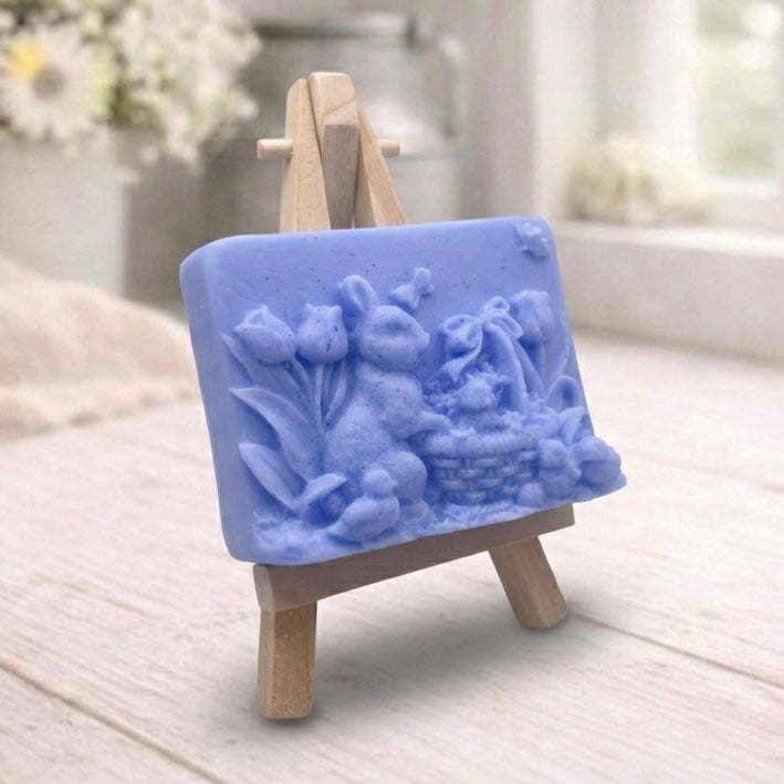 A rectangular handmade vegan soap in a sophisticated slate-grey tone, featuring a highly detailed relief scene of an Easter bunny standing next to a basket of eggs, surrounded by blooming tulips and small birds, displayed on a wooden easel.