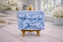 Load image into Gallery viewer, A rectangular handmade vegan soap in a sophisticated slate-grey tone, featuring a highly detailed relief scene of an Easter bunny standing next to a basket of eggs, surrounded by blooming tulips and small birds, displayed on a wooden easel.
