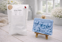 Load image into Gallery viewer, A rectangular handmade vegan soap in a sophisticated slate-grey tone, featuring a highly detailed relief scene of an Easter bunny standing next to a basket of eggs, surrounded by blooming tulips and small birds, displayed on a wooden easel.
