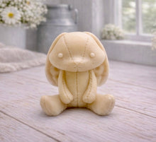 Load image into Gallery viewer, A handmade vegan soap sculpted in the shape of a sitting floppy-eared bunny rabbit with visible stitched seam details like a plush toy, in a soft yellow tone, displayed on a light wooden surface.
