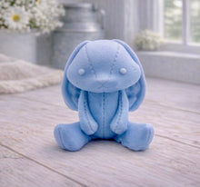 Load image into Gallery viewer, A handmade vegan soap sculpted in the shape of a sitting floppy-eared bunny rabbit with visible stitched seam details like a plush toy, in a soft blue tone, displayed on a light wooden surface.
