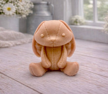 Load image into Gallery viewer, A handmade vegan soap sculpted in the shape of a sitting floppy-eared bunny rabbit with visible stitched seam details like a plush toy, in a soft peach-orange tone, displayed on a light wooden surface.

