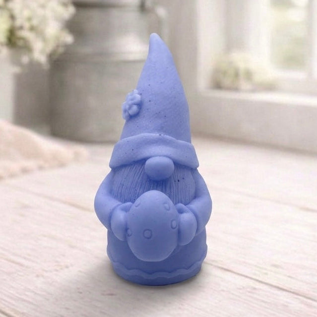 handmade vegan soap in the shape of a sitting gnome holding a decorated Easter egg, featuring a tall pointed hat with a flower detail and a textured beard, displayed on a neutral cloth in a bright spring setting.