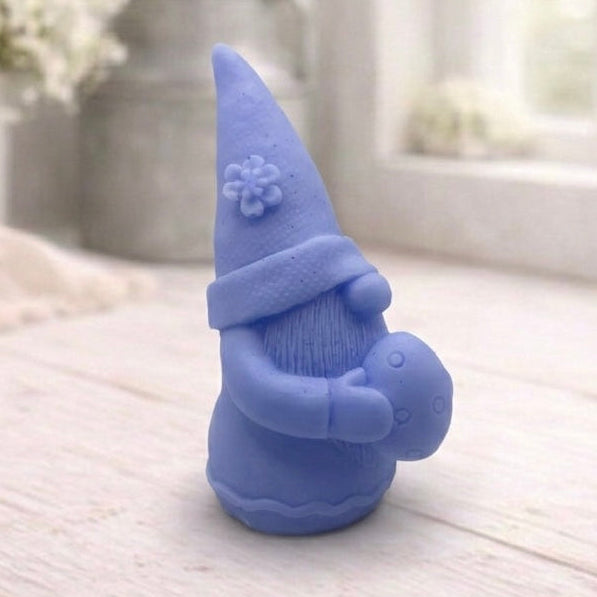 handmade vegan soap in the shape of a sitting gnome holding a decorated Easter egg, featuring a tall pointed hat with a flower detail and a textured beard, displayed on a neutral cloth in a bright spring setting.