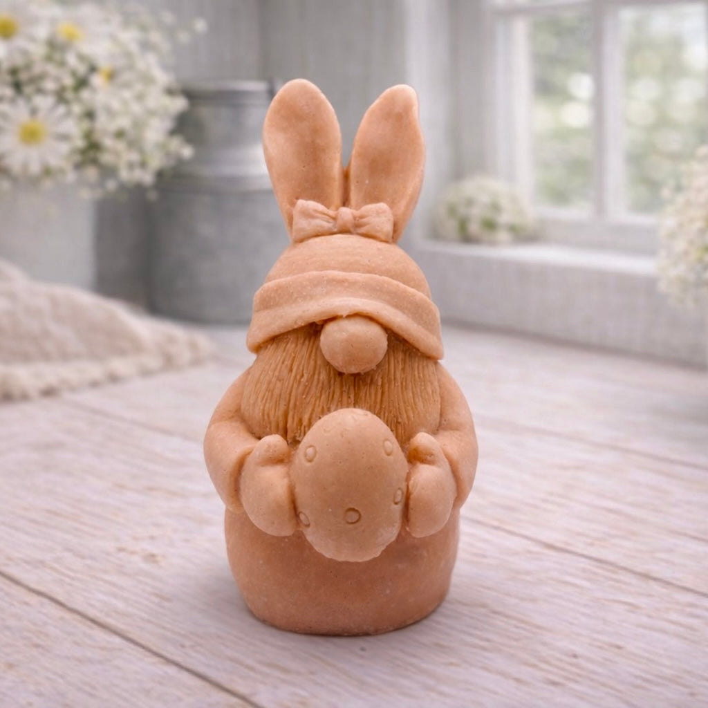 A detailed handmade vegan soap in a natural colour, sculpted as a whimsical Easter gnome (gonk) with long rabbit ears and a bow on its hat, holding a patterned Easter egg, displayed on a light wooden surface. Easter gift 