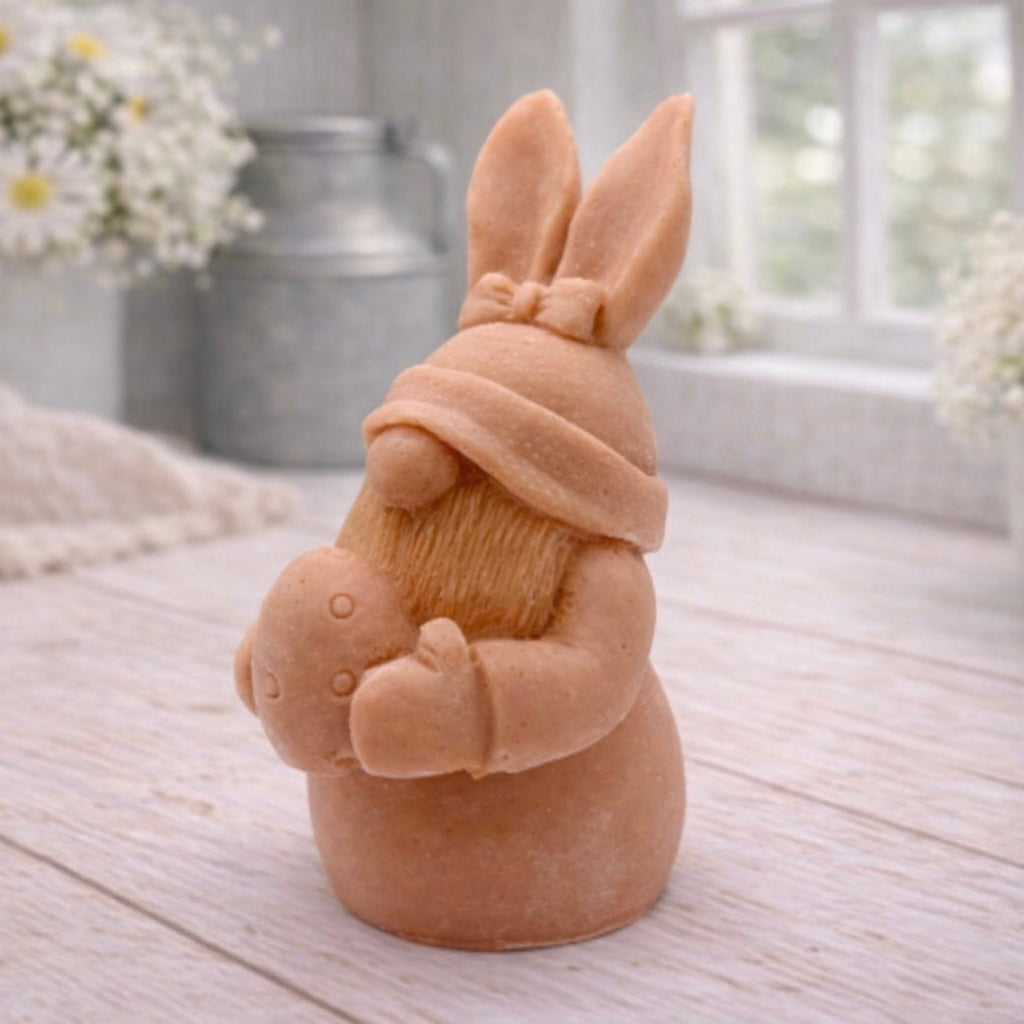 A detailed handmade vegan soap in a natural colour, sculpted as a whimsical Easter gnome (gonk) with long rabbit ears and a bow on its hat, holding a patterned Easter egg, displayed on a light wooden surface. Easter gift 