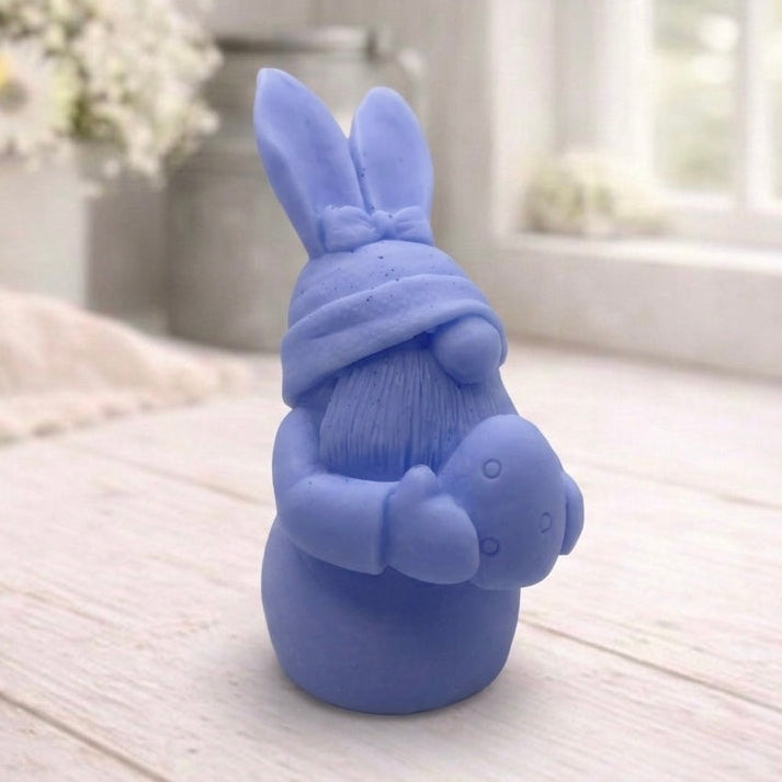 A detailed handmade vegan soap in a natural colour, sculpted as a whimsical Easter gnome (gonk) with long rabbit ears and a bow on its hat, holding a patterned Easter egg, displayed on a light wooden surface. Easter gift 