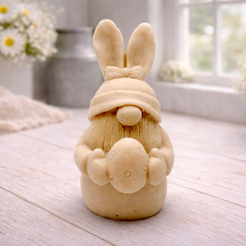 A detailed handmade vegan soap in a natural colour, sculpted as a whimsical Easter gnome (gonk) with long rabbit ears and a bow on its hat, holding a patterned Easter egg, displayed on a light wooden surface. Easter gift 
