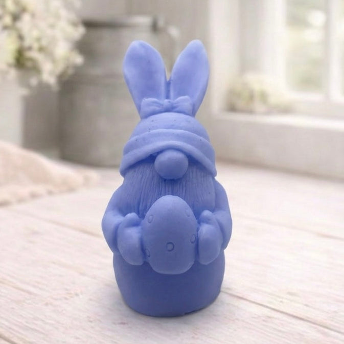 A detailed handmade vegan soap in a natural colour, sculpted as a whimsical Easter gnome (gonk) with long rabbit ears and a bow on its hat, holding a patterned Easter egg, displayed on a light wooden surface. Easter gift 