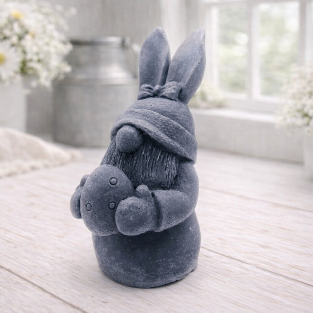A detailed handmade vegan soap in a natural colour, sculpted as a whimsical Easter gnome (gonk) with long rabbit ears and a bow on its hat, holding a patterned Easter egg, displayed on a light wooden surface.