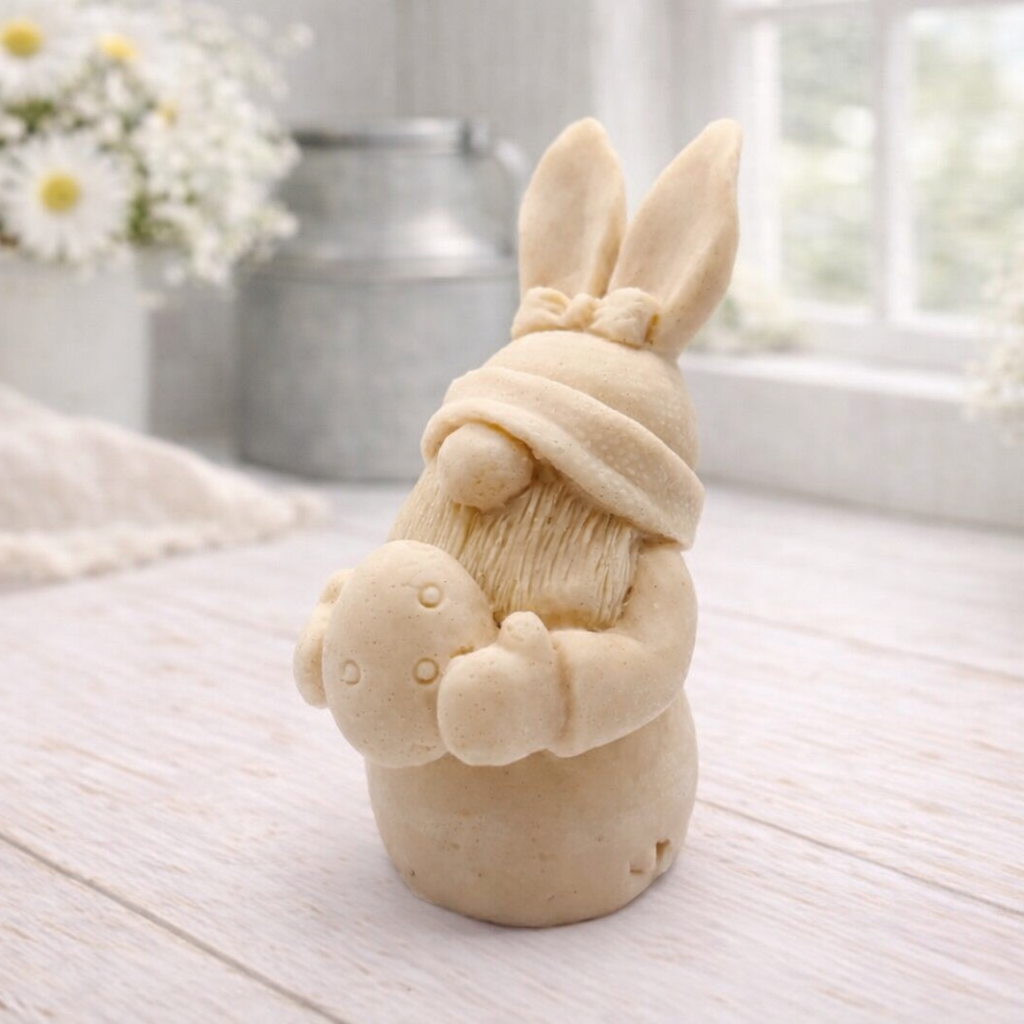 A detailed handmade vegan soap in a natural colour, sculpted as a whimsical Easter gnome (gonk) with long rabbit ears and a bow on its hat, holding a patterned Easter egg, displayed on a light wooden surface.