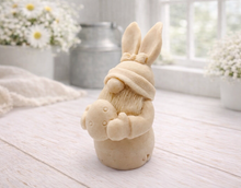 Load image into Gallery viewer, A detailed handmade vegan soap in a natural colour, sculpted as a whimsical Easter gnome (gonk) with long rabbit ears and a bow on its hat, holding a patterned Easter egg, displayed on a light wooden surface.

