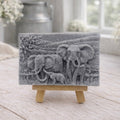 A rectangular handmade vegan soap in a charcoal grey tone, featuring a detailed relief carving of an elephant family—two adults and a small calf—walking through a textured savannah landscape, displayed on a miniature wooden easel.