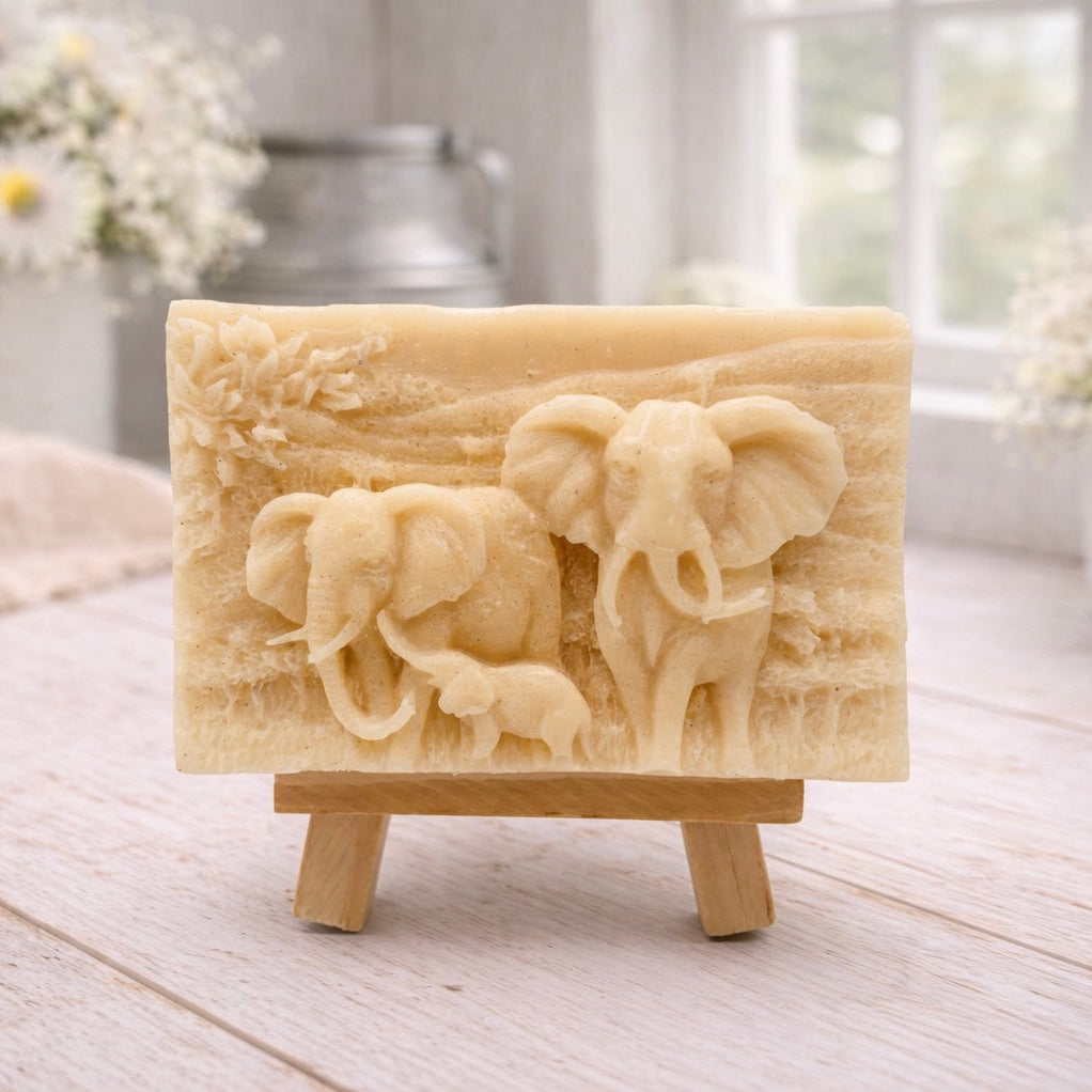 A rectangular handmade vegan soap in a warm yellow tone, featuring a detailed relief carving of an elephant family—two adults and a small calf—walking through a textured savannah landscape, displayed on a miniature wooden easel.
