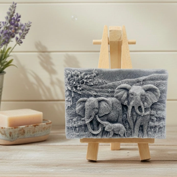 A rectangular handmade vegan soap, featuring a detailed relief carving of an elephant family—two adults and a small calf—walking through a textured savannah landscape, displayed on a miniature wooden easel.