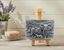 Load image into Gallery viewer, A rectangular handmade vegan soap, featuring a detailed relief carving of an elephant family—two adults and a small calf—walking through a textured savannah landscape, displayed on a miniature wooden easel.

