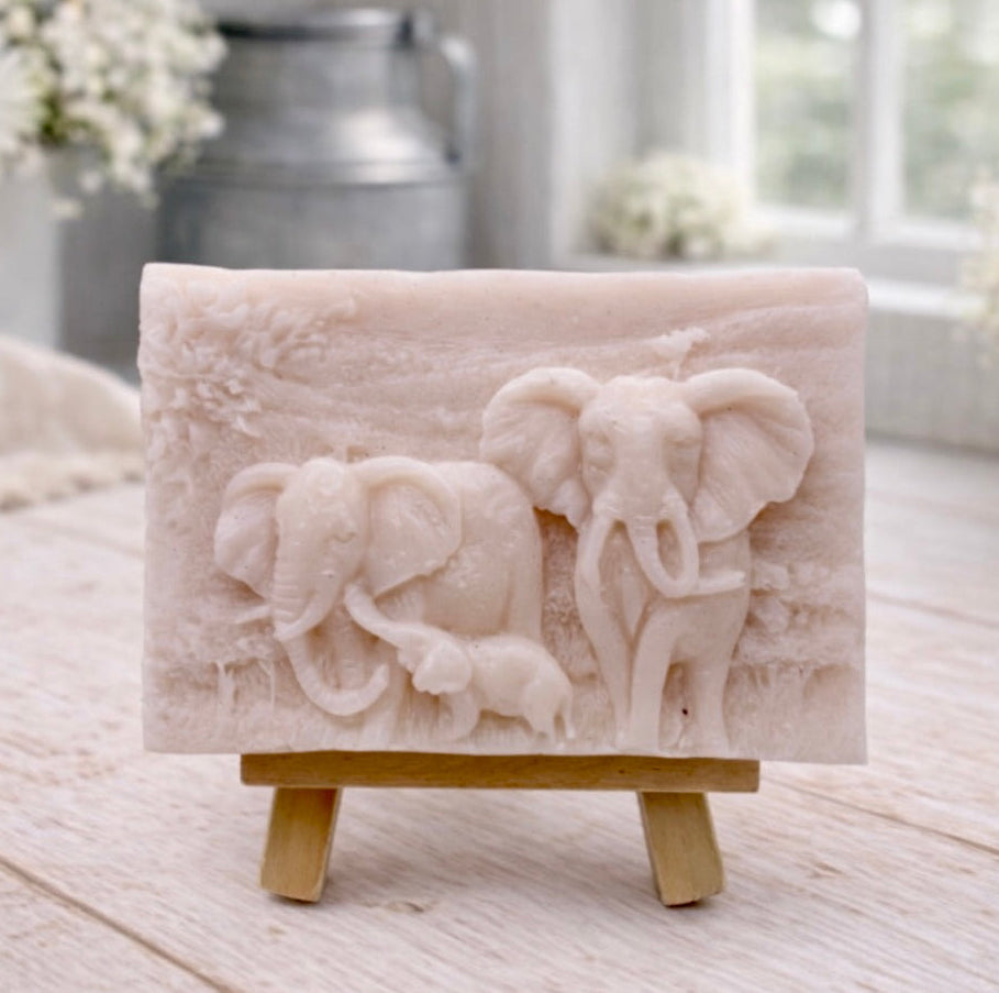 A rectangular handmade vegan soap, featuring a detailed relief carving of an elephant family—two adults and a small calf—walking through a textured savannah landscape, displayed on a miniature wooden easel.