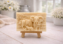 Load image into Gallery viewer, A rectangular handmade vegan soap in a warm yellow tone, featuring a detailed relief carving of an elephant family—two adults and a small calf—walking through a textured savannah landscape, displayed on a miniature wooden easel.

