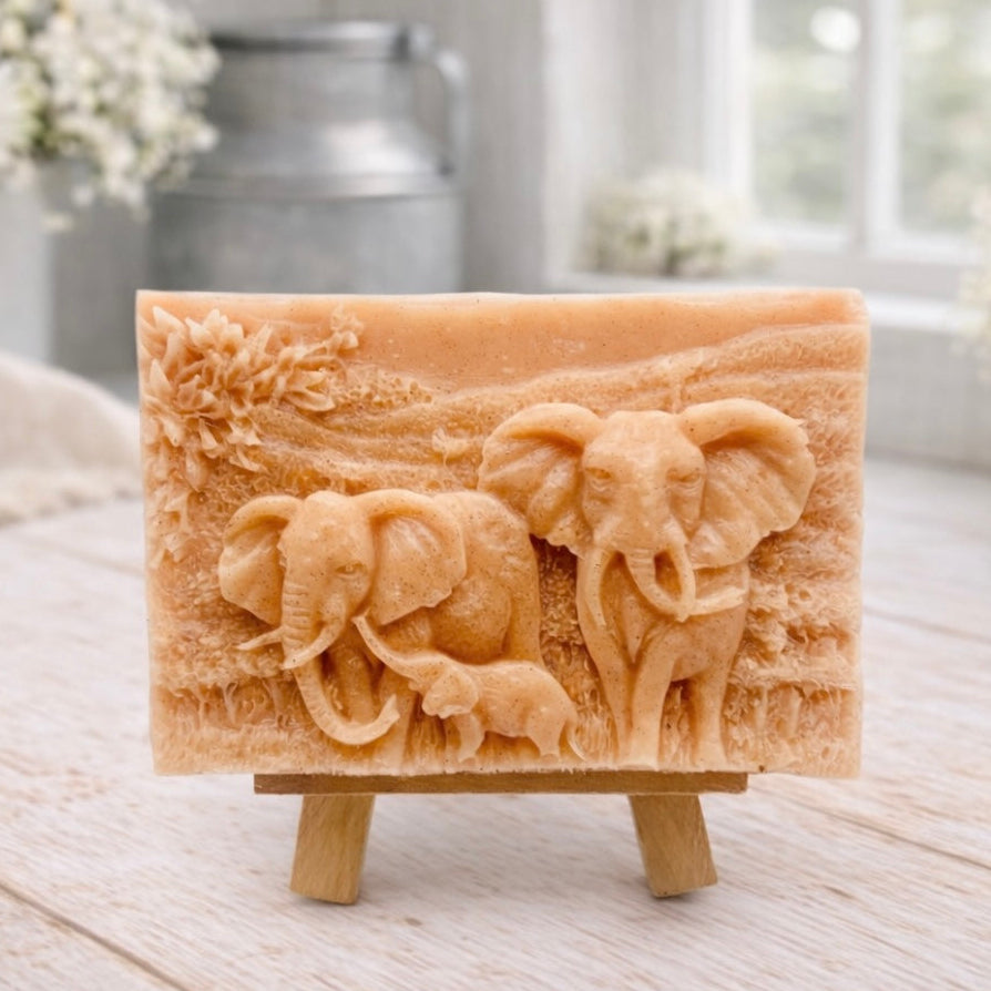 A rectangular handmade vegan soap in a warm terracotta tone, featuring a detailed relief carving of an elephant family—two adults and a small calf—walking through a textured savannah landscape, displayed on a miniature wooden easel.