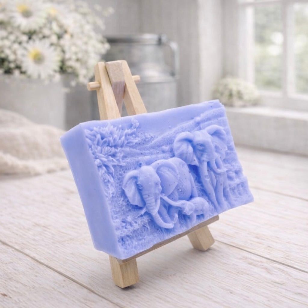 A rectangular handmade vegan soap in a warm blue tone, featuring a detailed relief carving of an elephant family—two adults and a small calf—walking through a textured savannah landscape, displayed on a miniature wooden easel.