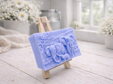 Load image into Gallery viewer, A rectangular handmade vegan soap in a warm blue tone, featuring a detailed relief carving of an elephant family—two adults and a small calf—walking through a textured savannah landscape, displayed on a miniature wooden easel.
