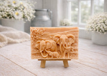 Load image into Gallery viewer, A rectangular handmade vegan soap in a warm terracotta tone, featuring a detailed relief carving of an elephant family—two adults and a small calf—walking through a textured savannah landscape, displayed on a miniature wooden easel.
