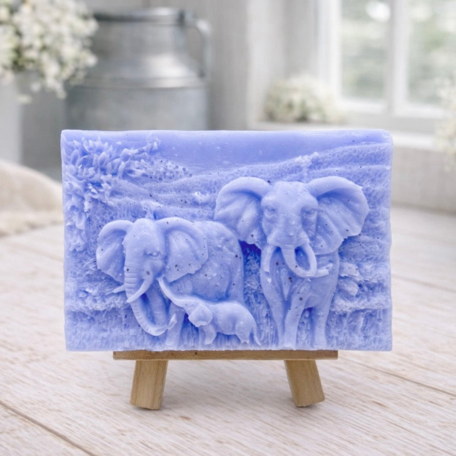 A rectangular handmade vegan soap, featuring a detailed relief carving of an elephant family—two adults and a small calf—walking through a textured savannah landscape, displayed on a miniature wooden easel.