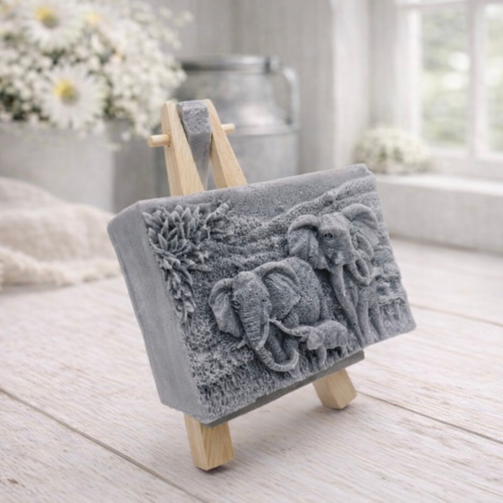 A rectangular handmade vegan soap in a grey tone, featuring a detailed relief carving of an elephant family—two adults and a small calf—walking through a textured savannah landscape, displayed on a miniature wooden easel.
