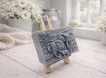 Load image into Gallery viewer, A rectangular handmade vegan soap in a grey tone, featuring a detailed relief carving of an elephant family—two adults and a small calf—walking through a textured savannah landscape, displayed on a miniature wooden easel.
