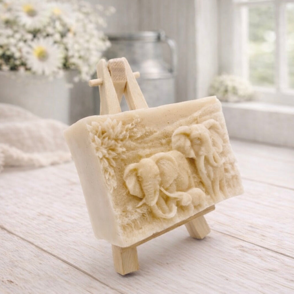 A rectangular handmade vegan soap in a warm yellow tone, featuring a detailed relief carving of an elephant family—two adults and a small calf—walking through a textured savannah landscape, displayed on a miniature wooden easel.