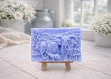 Load image into Gallery viewer, A rectangular handmade vegan soap, featuring a detailed relief carving of an elephant family—two adults and a small calf—walking through a textured savannah landscape, displayed on a miniature wooden easel.
