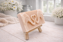 Load image into Gallery viewer, A rectangular handmade vegan soap  featuring a detailed relief of two elephants facing each other with their trunks entwined to form a heart shape, displayed on a miniature wooden easel.
