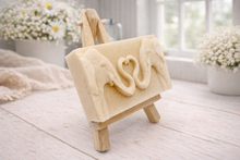 Load image into Gallery viewer, A rectangular handmade vegan soap  featuring a detailed relief of two elephants facing each other with their trunks entwined to form a heart shape, displayed on a miniature wooden easel.
