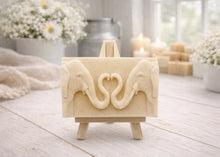 Load image into Gallery viewer, A rectangular handmade vegan soap  featuring a detailed relief of two elephants facing each other with their trunks entwined to form a heart shape, displayed on a miniature wooden easel.
