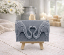 Load image into Gallery viewer, A rectangular handmade vegan soap in a slate-grey tone, featuring a detailed relief of two elephants facing each other with their trunks entwined to form a heart shape, displayed on a miniature wooden easel.
