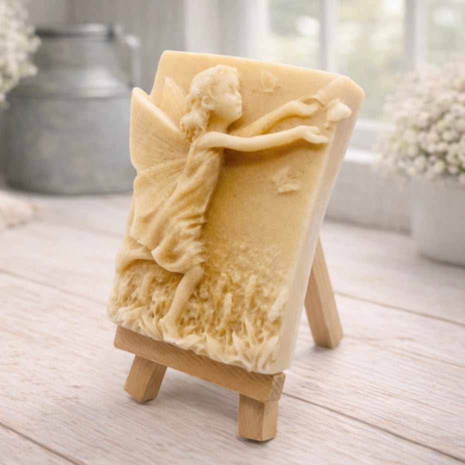 A rectangular handmade vegan soap featuring a detailed relief of a winged fairy reaching towards a butterfly in a meadow of tall grass and flowers, displayed on a miniature wooden easel