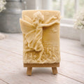 A rectangular handmade vegan soap featuring a detailed relief of a winged fairy reaching towards a butterfly in a meadow of tall grass and flowers, displayed on a miniature wooden easel
