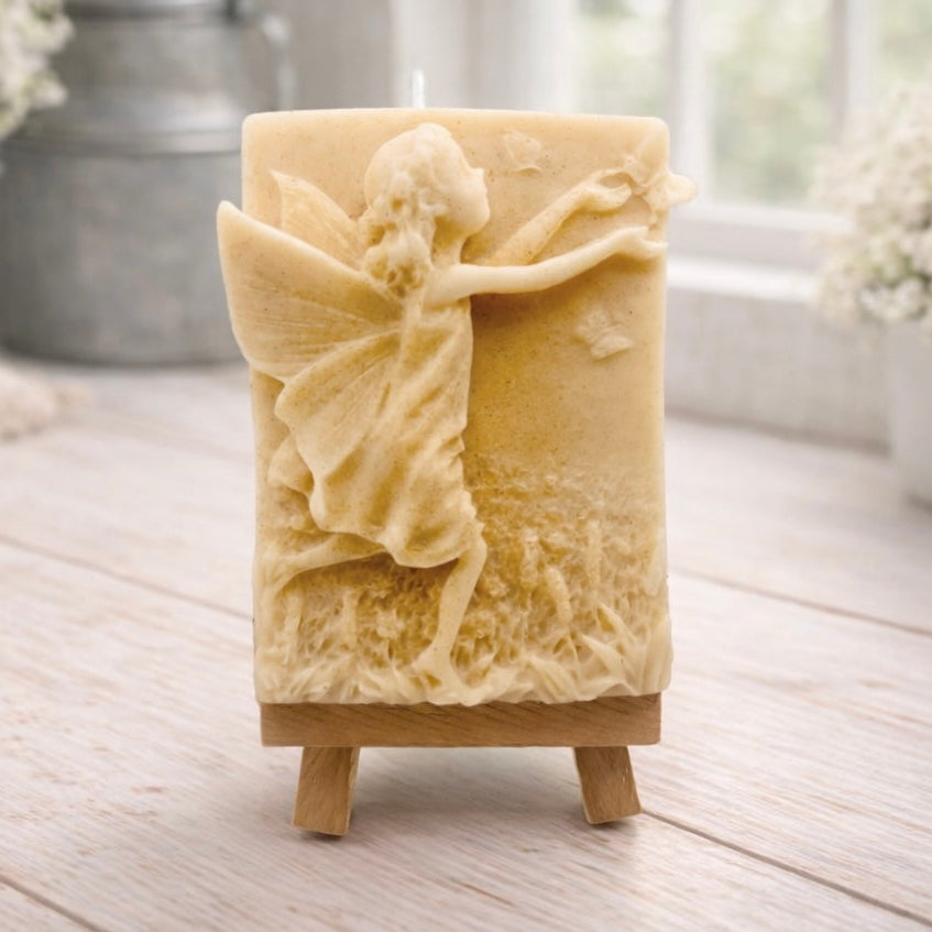A rectangular handmade vegan soap featuring a detailed relief of a winged fairy reaching towards a butterfly in a meadow of tall grass and flowers, displayed on a miniature wooden easel