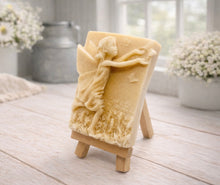 Load image into Gallery viewer, A rectangular handmade vegan soap featuring a detailed relief of a winged fairy reaching towards a butterfly in a meadow of tall grass and flowers, displayed on a miniature wooden easel
