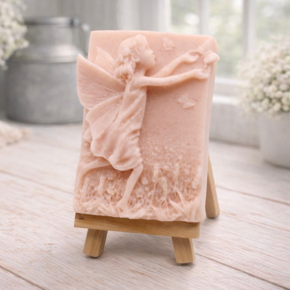 A rectangular handmade vegan soap featuring a detailed relief of a winged fairy reaching towards a butterfly in a meadow of tall grass and flowers, displayed on a miniature wooden easel