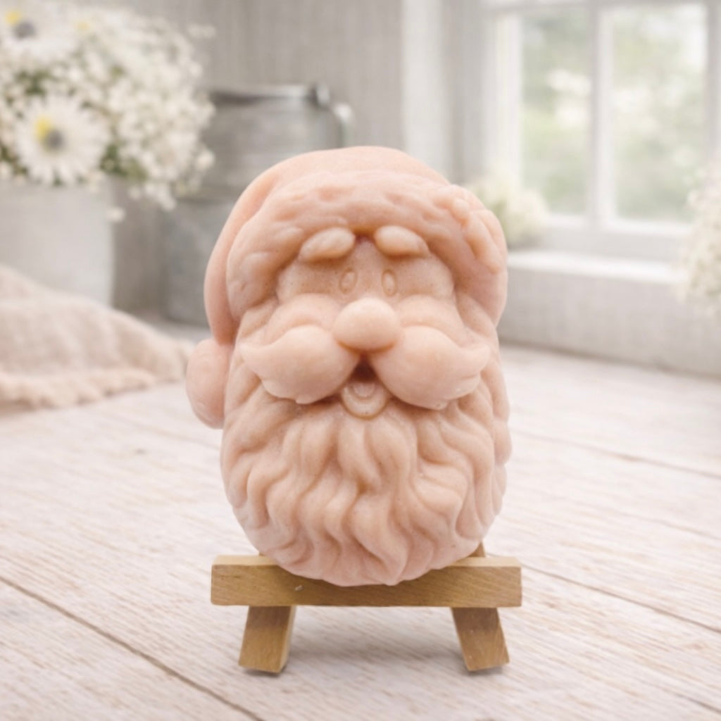 handmade vegan soap sculpted in the shape of Father Christmas's head, featuring a detailed curly beard, a jolly moustache, and a classic Santa hat, displayed on a miniature wooden easel in a bright farmhouse setting.
