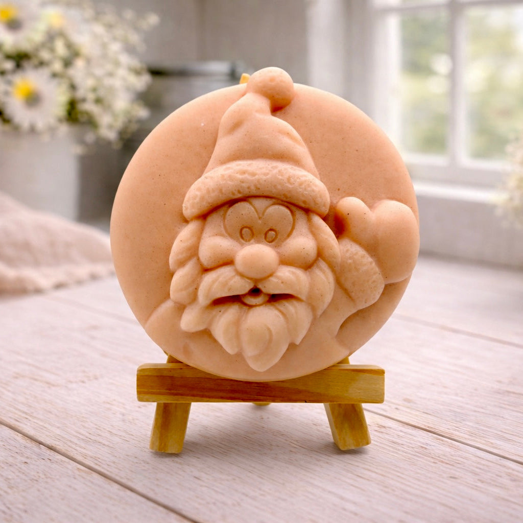 A circular handmade vegan soap featuring a detailed high-relief carving of a jolly Father Christmas waving, with a textured beard and a classic Santa hat, displayed on a miniature wooden easel.