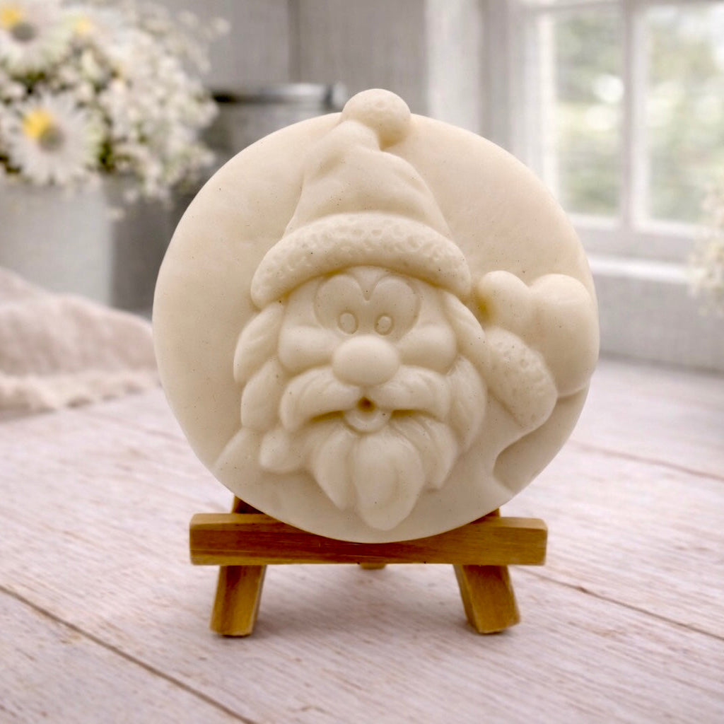 A circular handmade vegan soap featuring a detailed high-relief carving of a jolly Father Christmas waving, with a textured beard and a classic Santa hat, displayed on a miniature wooden easel.